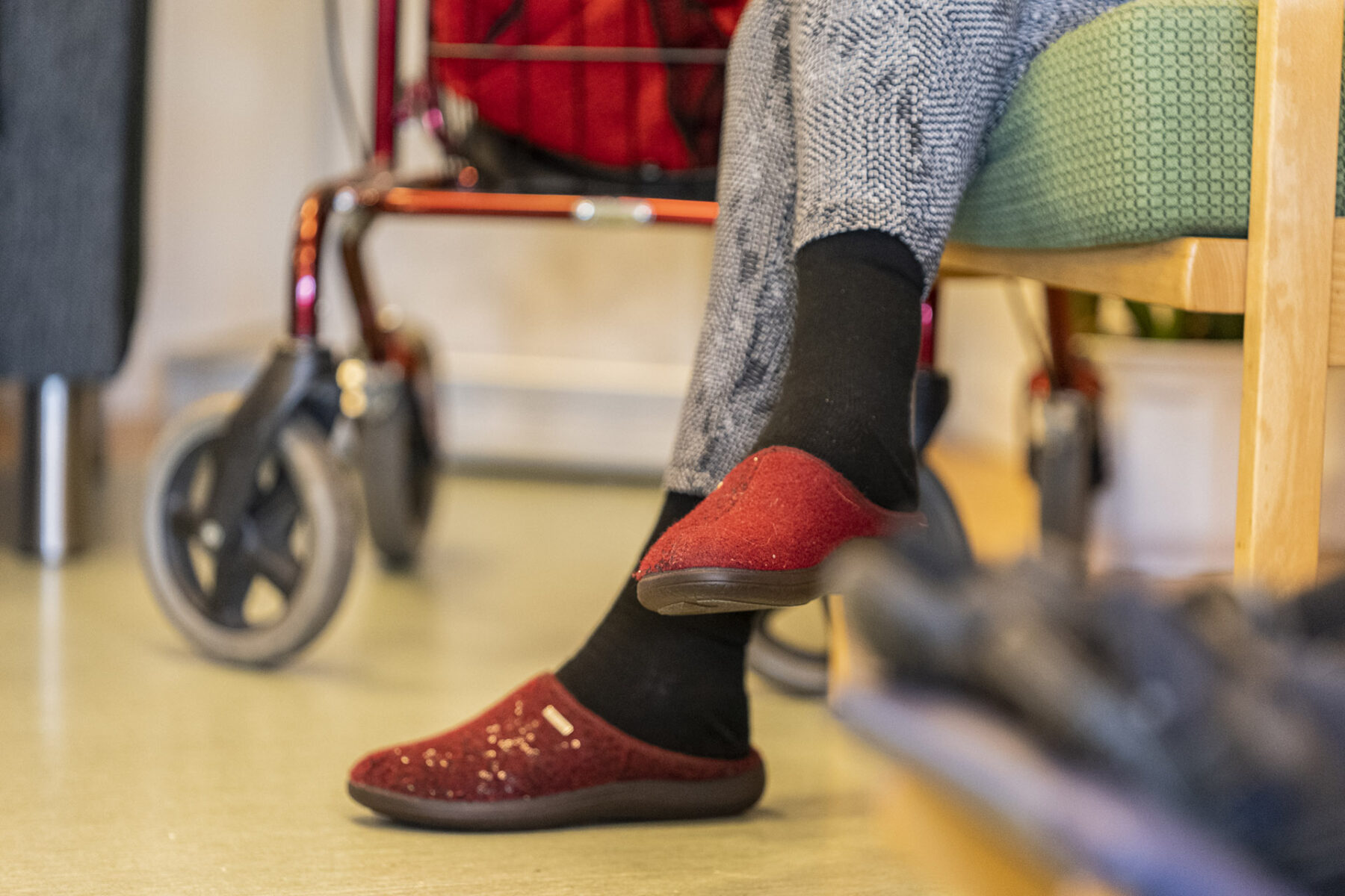 Närbild på äldre persons toffelbeklädda fötter framför en suddig rullator.